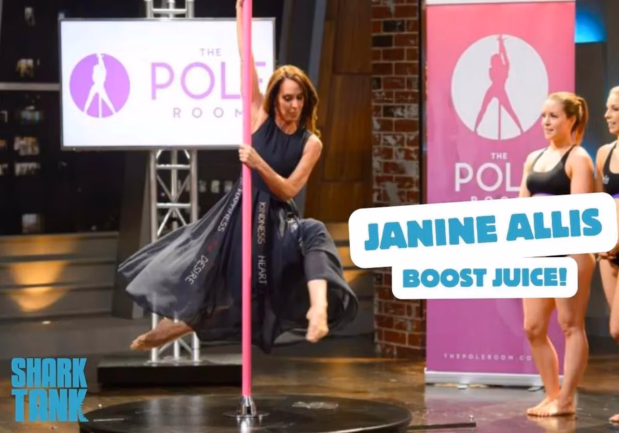 Woman performing a pole dancing move on a pink pole wearing a black dress, with two women in black athletic wear looking on, Shark Tank logo in bottom left, and The Pole Room banners in background.