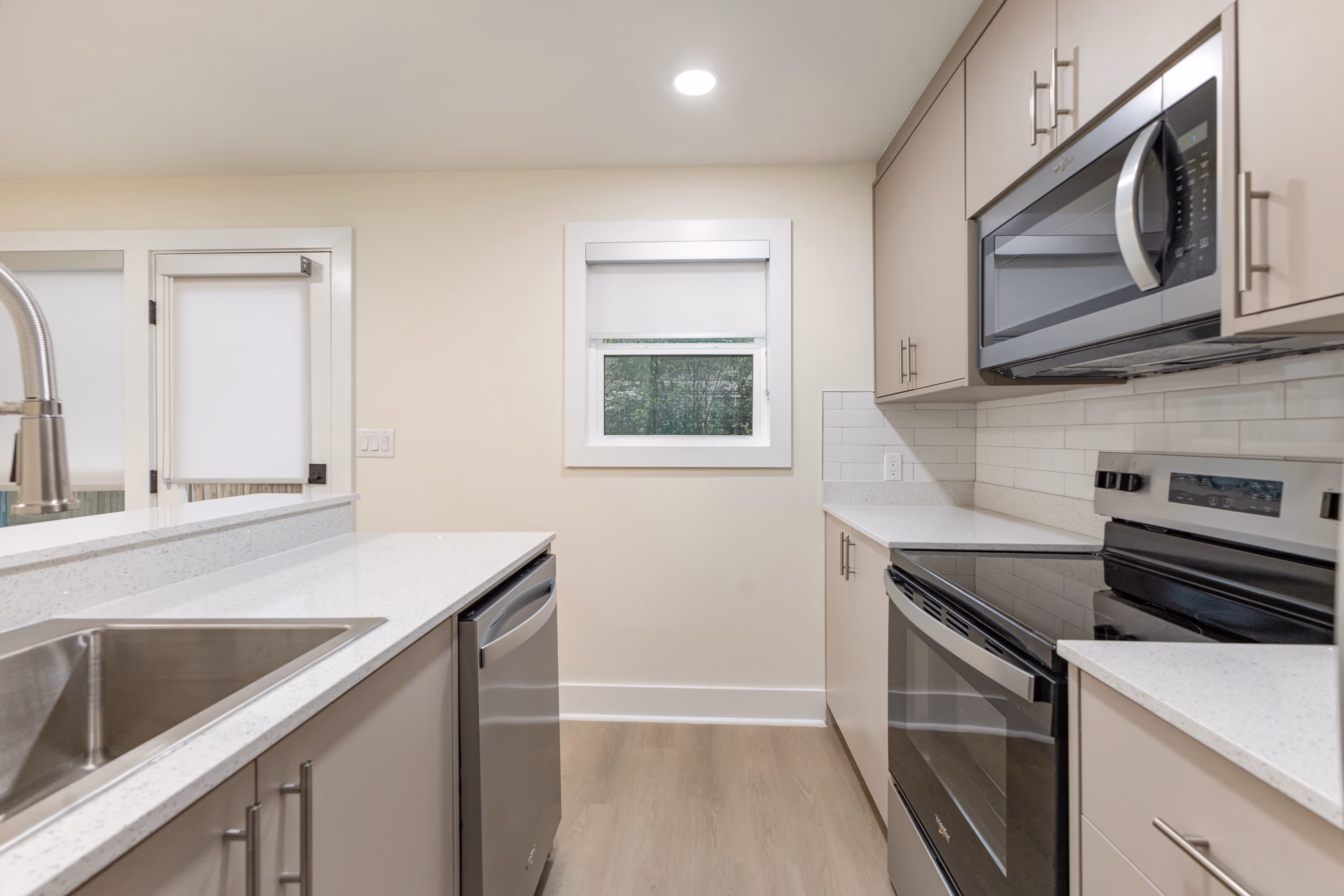 Modern kitchen with light beige cabinets, stainless steel appliances including stove, microwave, and dishwasher, white countertops, and a small window above the countertop.