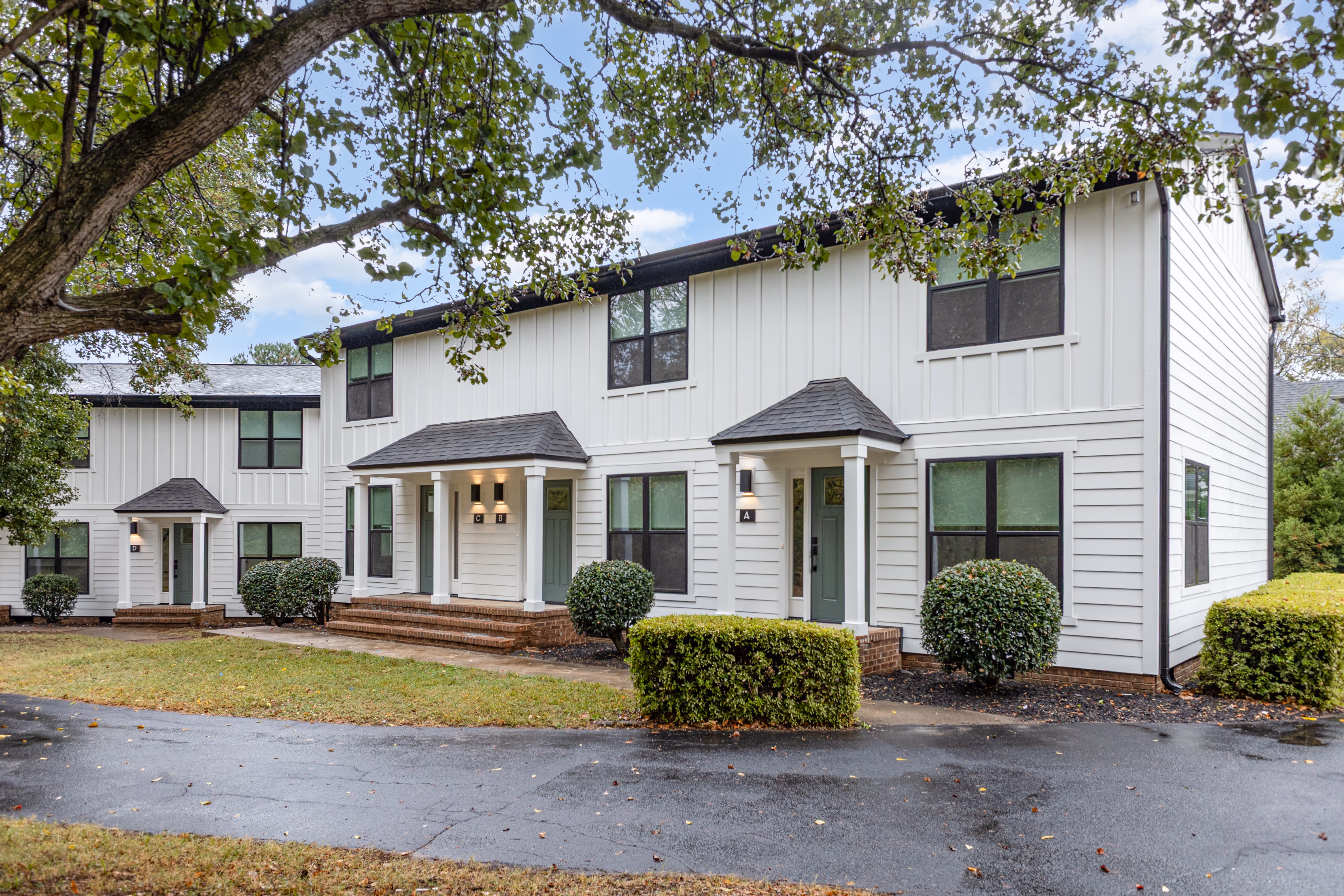 White two-story townhouse with black window frames, green doors, brick steps, and trimmed bushes in front.