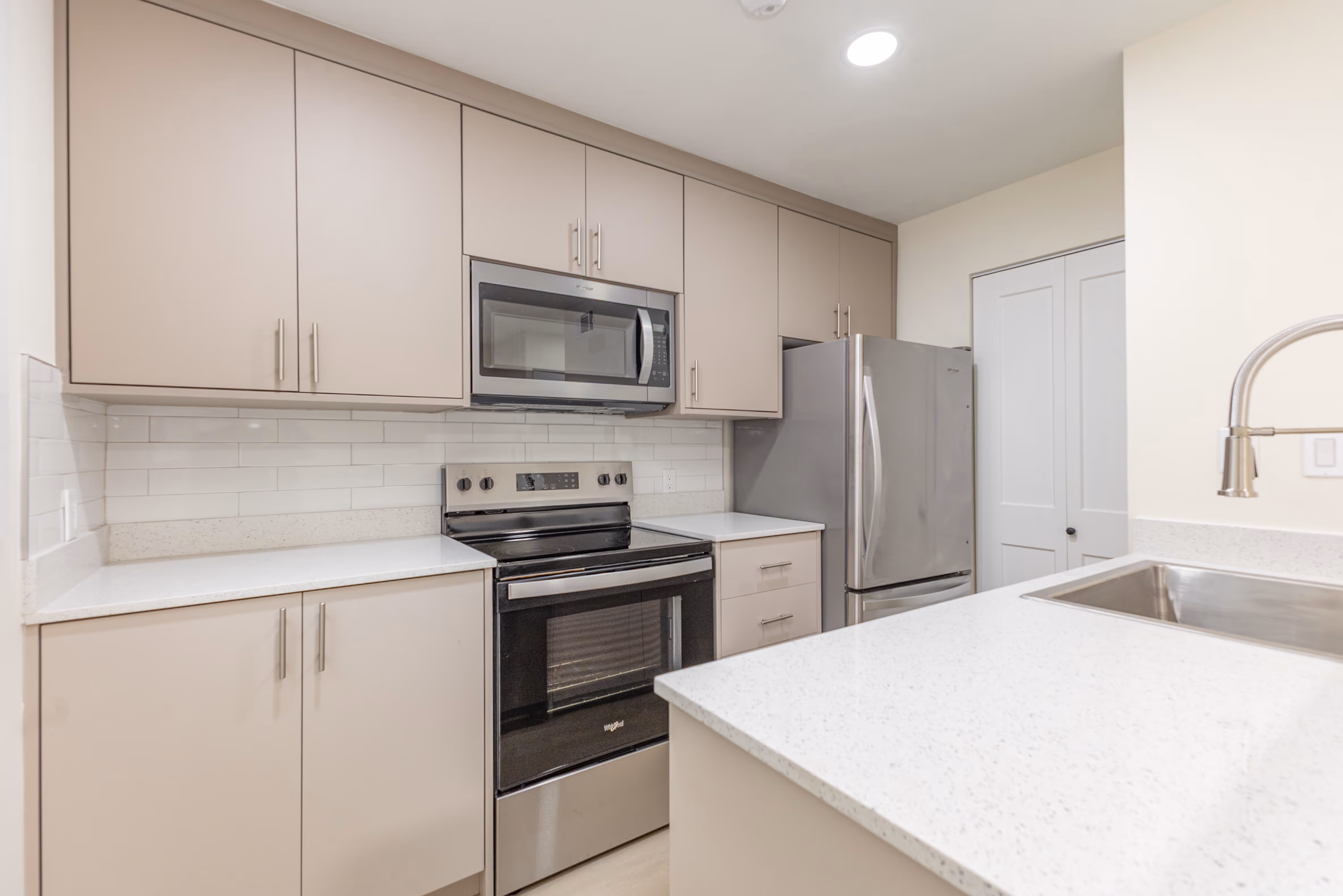 Modern kitchen with beige cabinets, stainless steel microwave, oven, refrigerator, white tiled backsplash, and quartz countertops.