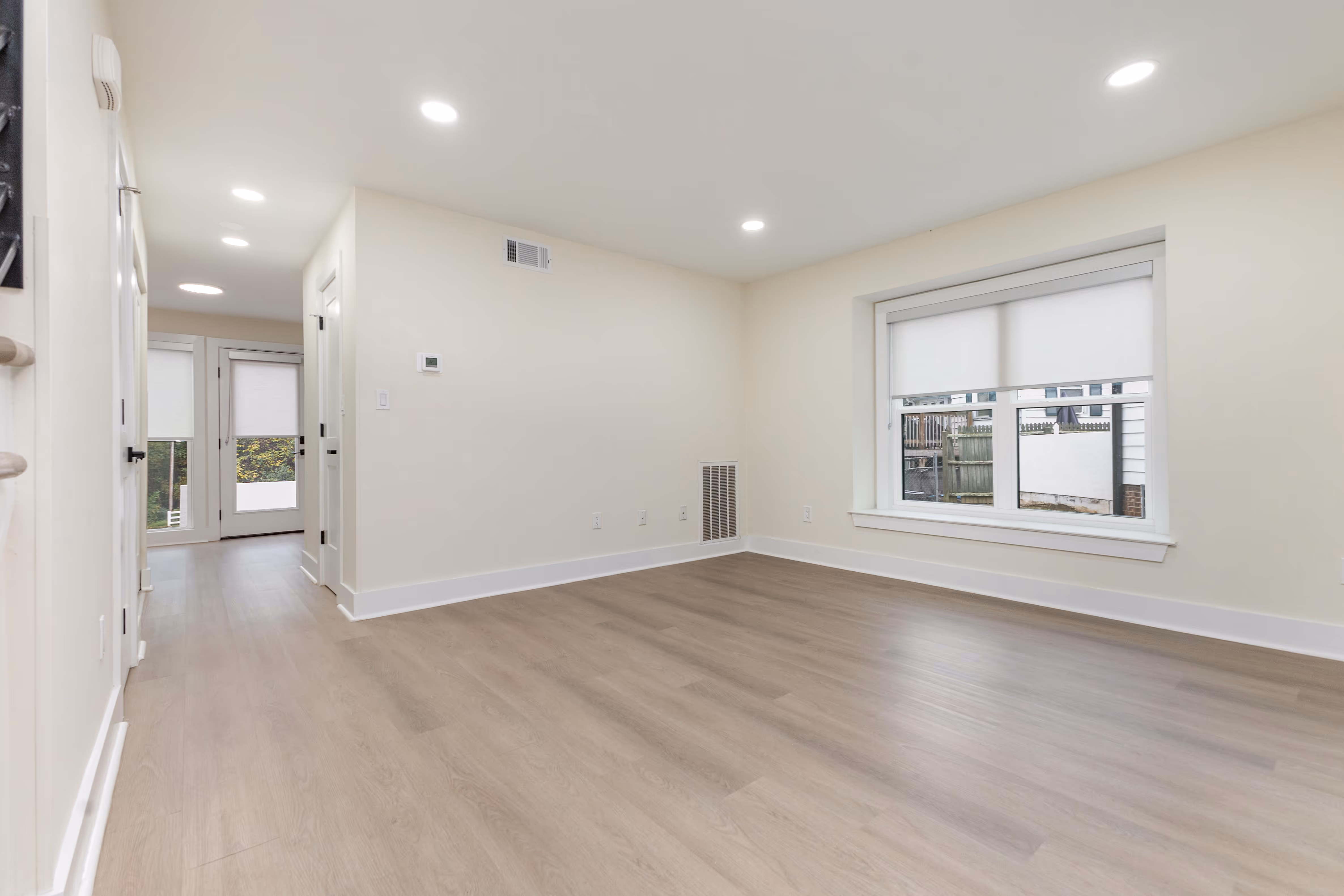 Empty room with light wood flooring, beige walls, recessed ceiling lights, and a window with white roller shades showing an outdoor fence.