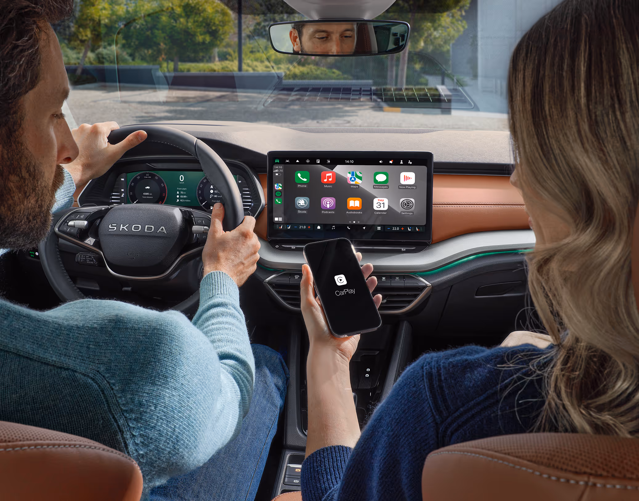 Blick von der Rückbank in das Cockpit des Škoda Octavia Balance, in dem Fahrer und Beifahrerin zu sehen sind. Die Beifahrerin hält ein Smartphone, das via Wireless SmartLink mit dem Infotainment-System verbunden ist, auf dessen Display die Apple CarPlay Oberfläche zu sehen ist. Die Szene verdeutlicht die Konnektivitäts-Features des Fahrzeugs vor einer unscharfen urbanen Kulisse im Hintergrund.