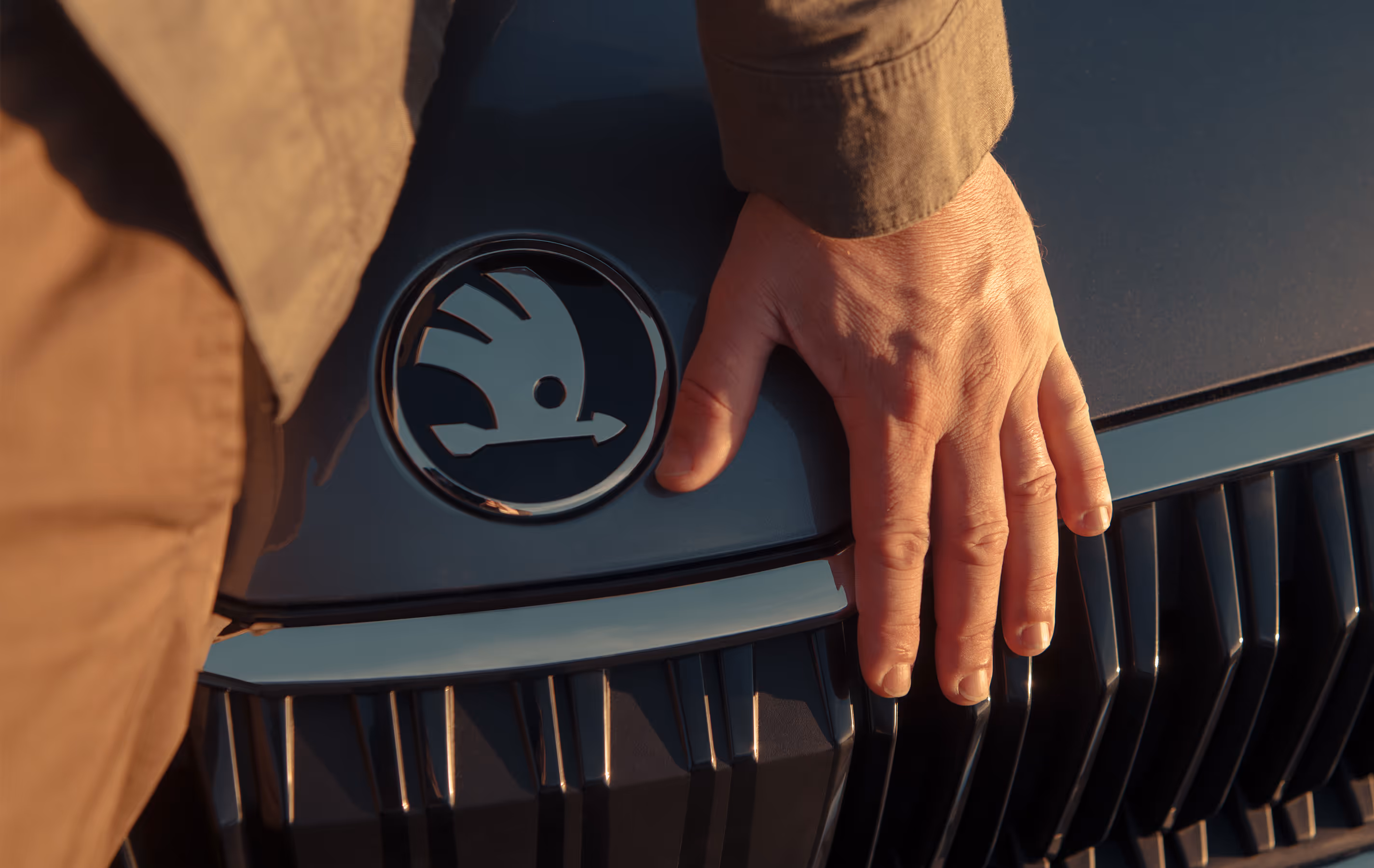 Eine Nahaufnahme zeigt die Hand eines Mannes, die sanft auf der Motorhaube des Škoda Octavia Balance ruht, direkt neben dem markanten Škoda Logo. Das Bild fokussiert auf die Design-Details der Frontpartie und den Kühlergrill, die von warmem, goldenem Sonnenlicht angestrahlt werden.