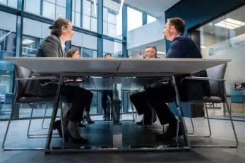 Vier Personen sitzen an einem Konferenztisch in einem modernen Bürogebäude und führen eine Besprechung.