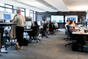 Büroumgebung mit mehreren Personen, die an Schreibtischen mit Computern arbeiten.