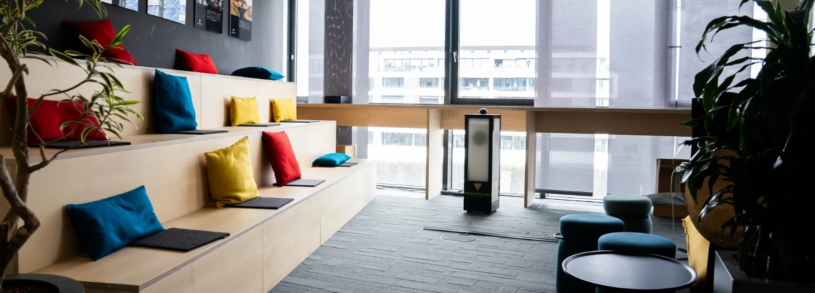 Moderner Besprechungsraum mit abgestuften Holzsitzen, bunten Kissen und großen Fenstern mit halbtransparenten Rollos.