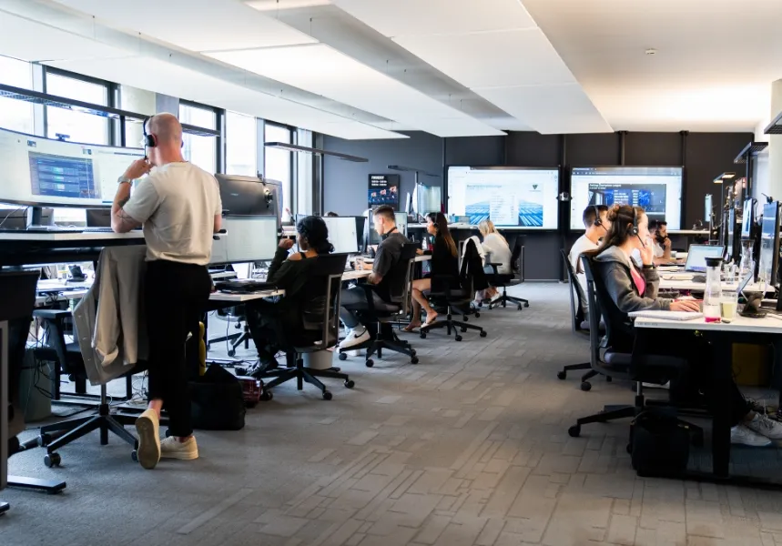 Moderne Büroarbeitsplätze mit mehreren Personen, die an stehenden und sitzenden Schreibtischen arbeiten, umgeben von Monitoren und Computern.