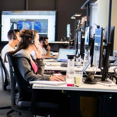 Drei Personen mit Headsets arbeiten an Computern in einem modernen Büro mit einem großen Bildschirm im Hintergrund.