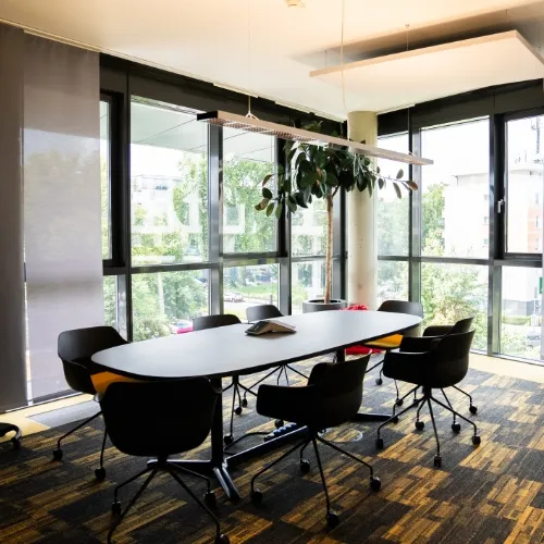 Moderner Konferenzraum mit ovalem Tisch, sechs schwarzen Bürostühlen auf Rollen und großen Fenstern mit Blick ins Grüne.