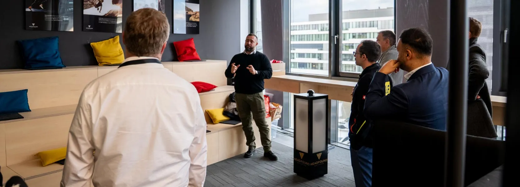 Ein Mann spricht vor einer kleinen Gruppe in einem modernen Besprechungsraum mit mehrstöckiger Sitzbank und bunten Kissen.