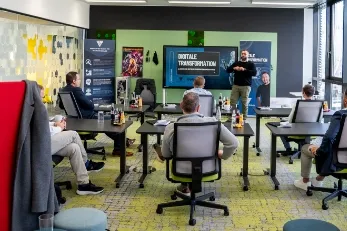 Büroszene mit mehreren Personen in einem Meetingraum, ein Mann präsentiert vor einem Monitor mit der Aufschrift 'Digitale Transformation'.