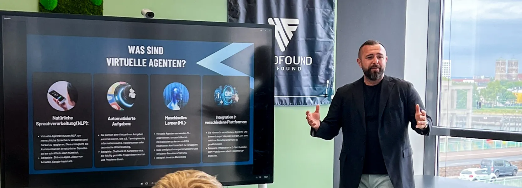 Ein Mann gibt einen Vortrag vor einem Bildschirm mit der Überschrift 'Was sind virtuelle Agenten?' und vier Informationsfeldern zu Sprachverarbeitung, automatisierten Aufgaben, maschinellem Lernen und Plattformintegration.