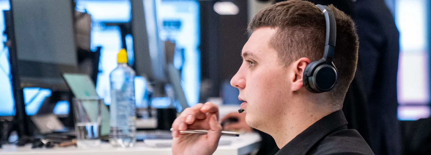 Ein junger Mann mit Headset sitzt am Schreibtisch und hält einen Stift, vor ihm sind unscharfe Computerbildschirme.