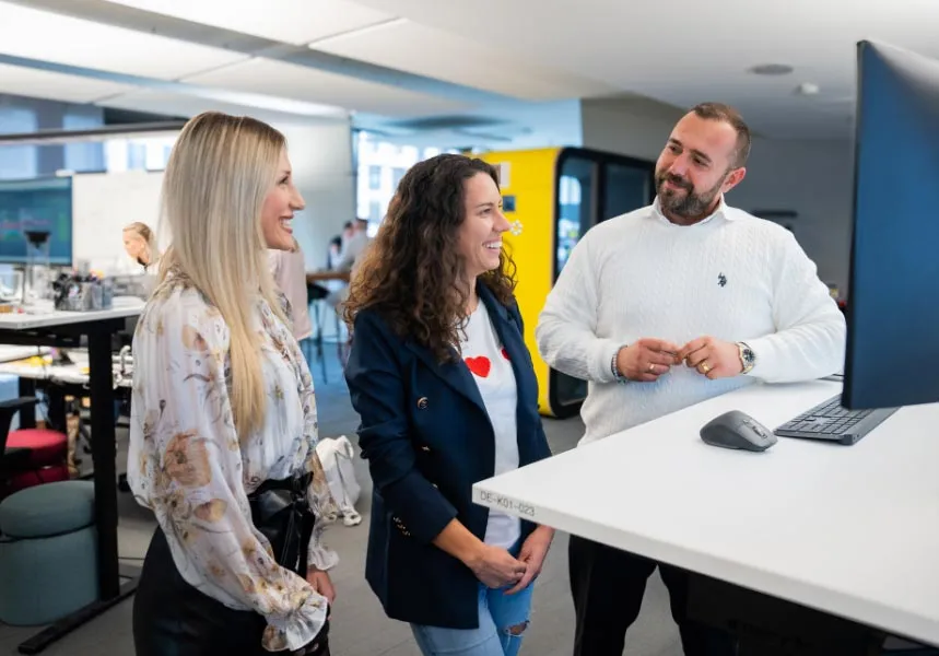 Drei Personen unterhalten sich lachend in einem modernen Büro vor einem Computerbildschirm.