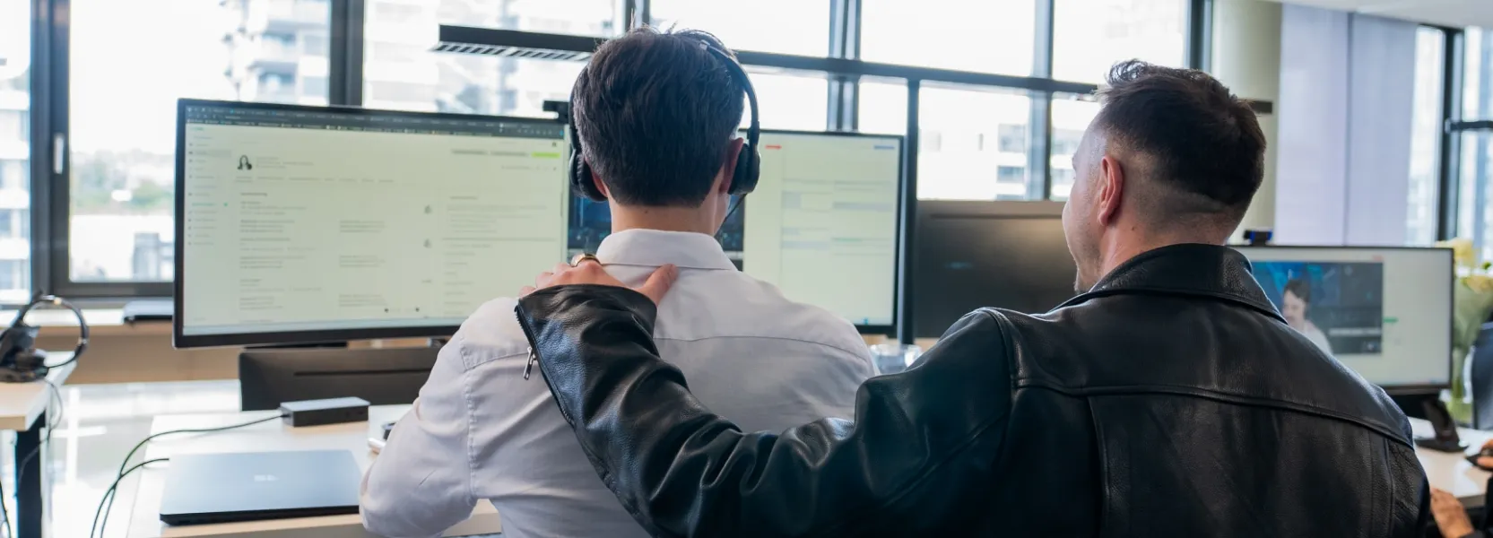 Zwei Männer am Büroarbeitsplatz, einer sitzt mit Headset vor zwei Monitoren, der andere legt unterstützend die Hand auf seine Schulter.