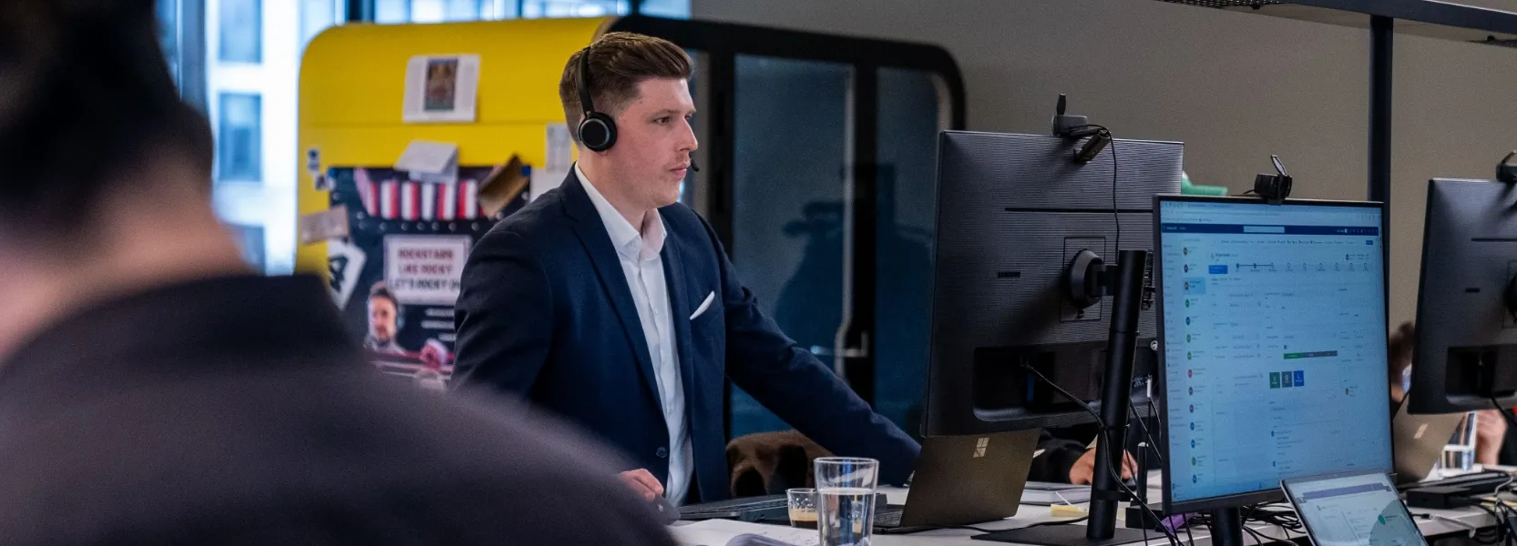 Geschäftsmann mit Headset arbeitet an einem Stehtisch vor mehreren Monitoren in einem modernen Büro.