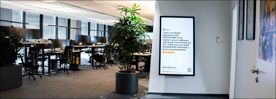 Modernes Büro mit mehreren Schreibtischen, Computern und einem großen Pflanzenkübel neben einem digitalen Display mit Kundenbewertung.