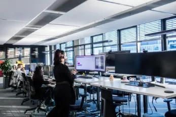 Büroumgebung mit mehreren Personen, die an Schreibtischen mit Computern arbeiten.