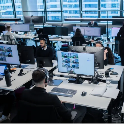 Drei Personen mit Headsets arbeiten an Computern in einem modernen Büro mit einem großen Bildschirm im Hintergrund.