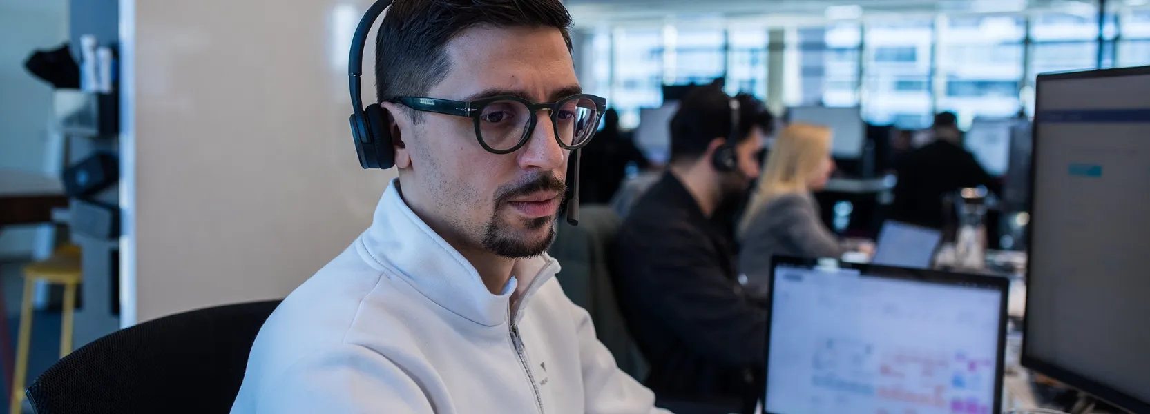 Ein junger Mann mit Headset sitzt am Schreibtisch und hält einen Stift, vor ihm sind unscharfe Computerbildschirme.