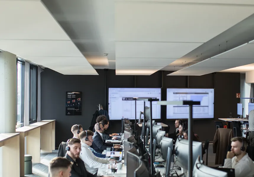 Moderne Büroarbeitsplätze mit mehreren Personen, die an stehenden und sitzenden Schreibtischen arbeiten, umgeben von Monitoren und Computern.