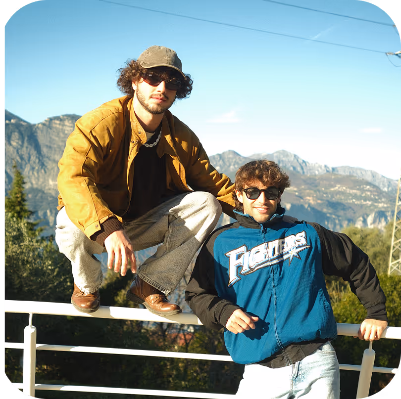 Two young men wearing sunglasses posing outdoors on a railing with mountains and clear sky in the background.