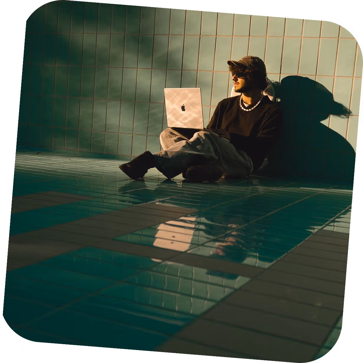 Man wearing a cap and sunglasses sitting against a tiled wall with a laptop on his lap, lit by warm sunlight.