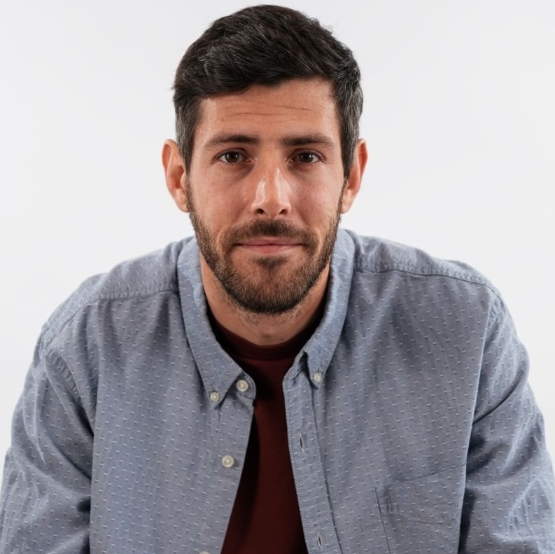 Man with dark hair and beard wearing a textured light blue button-up shirt over a maroon t-shirt against a plain white background.