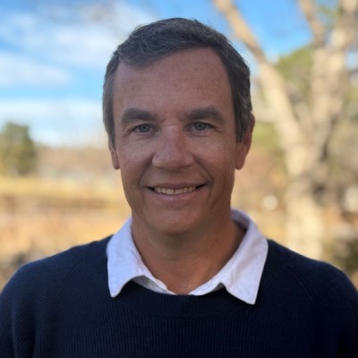 Smiling man with short dark hair wearing a dark sweater over a white collared shirt outdoors with blurred natural background.
