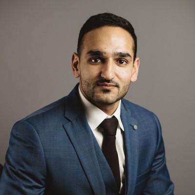 Man with short dark hair wearing a blue suit, white shirt, and dark tie, posing against a plain gray background.