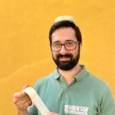 Man with glasses and beard smiling, wearing a green shirt with a name tag reading 'Giulio', holding a roll of tape and balancing a piece on his head against a yellow background.