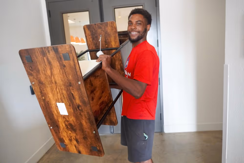 Cheerful professional mover carrying a disassembled wooden table down a hallway in Austin, TX, demonstrating careful residential moving service.
