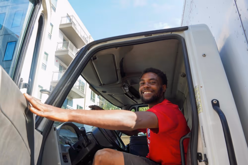 Happy, bearded Austin TX moving company driver in a red shirt sitting in the cab of a commercial moving truck, ready to drive for an interstate move.