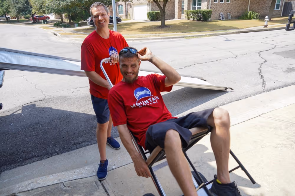 Two happy professional Austin TX movers posing with a dolly and moving truck ramp in a sunny residential neighborhood.