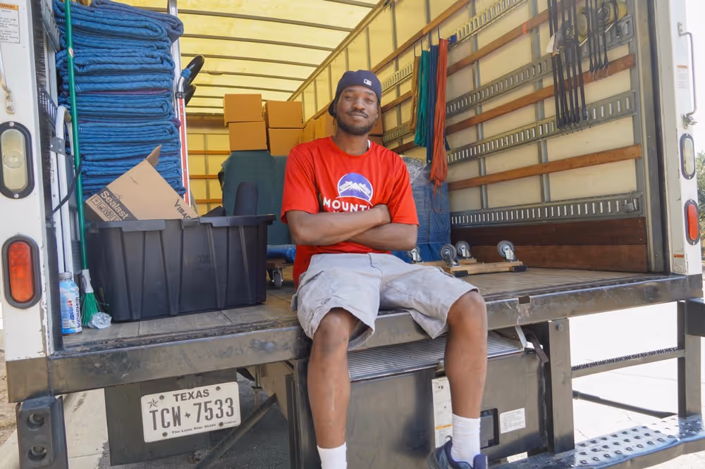 Austin TX mover taking a break, sitting on the tailgate of a moving truck loaded with boxes, blankets, and supplies in Texas.