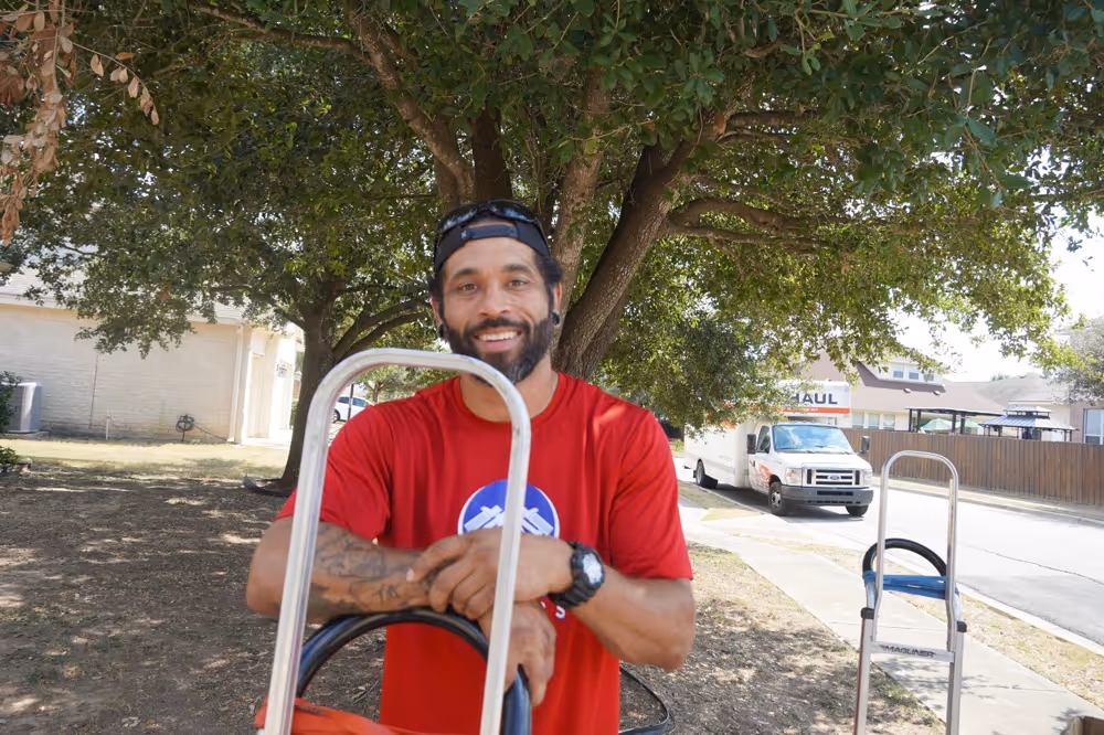 Friendly Austin TX mover in red shirt holding hand truck under large tree with U-Haul truck visible on residential street.