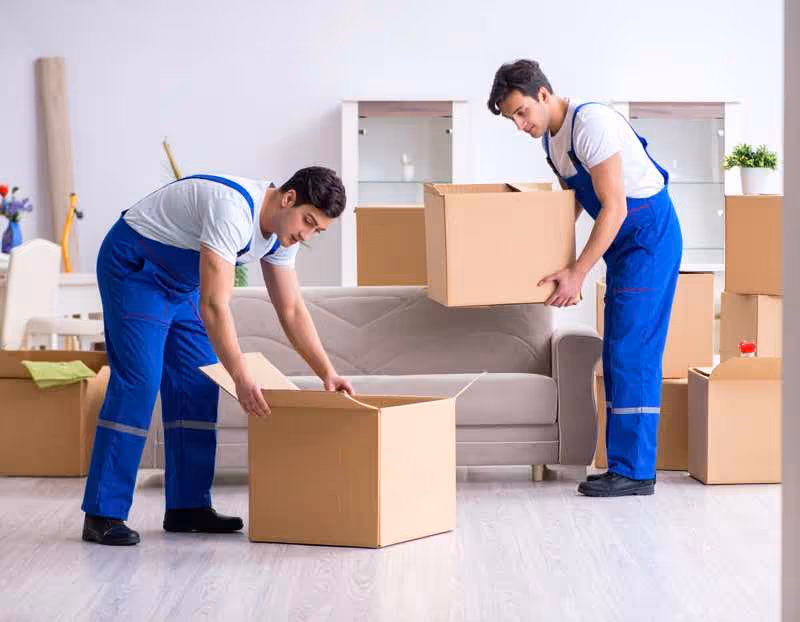 Two uniformed movers wearing blue overalls lifting and preparing to pack a large cardboard box inside an office/apartment. Keywords: Austin TX movers, professional moving help, safe lifting.