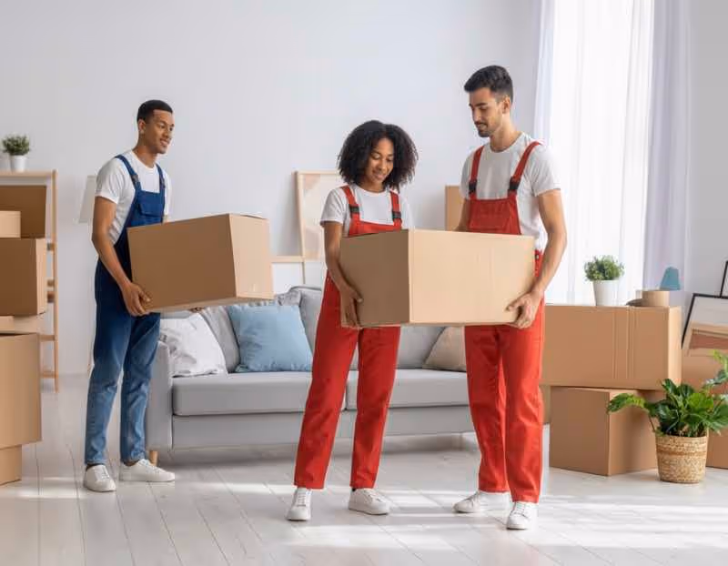 Diverse moving team in red and blue overalls carrying large moving boxes in a bright, modern living space. Keywords: local movers Austin, furniture and box moving, residential relocation.