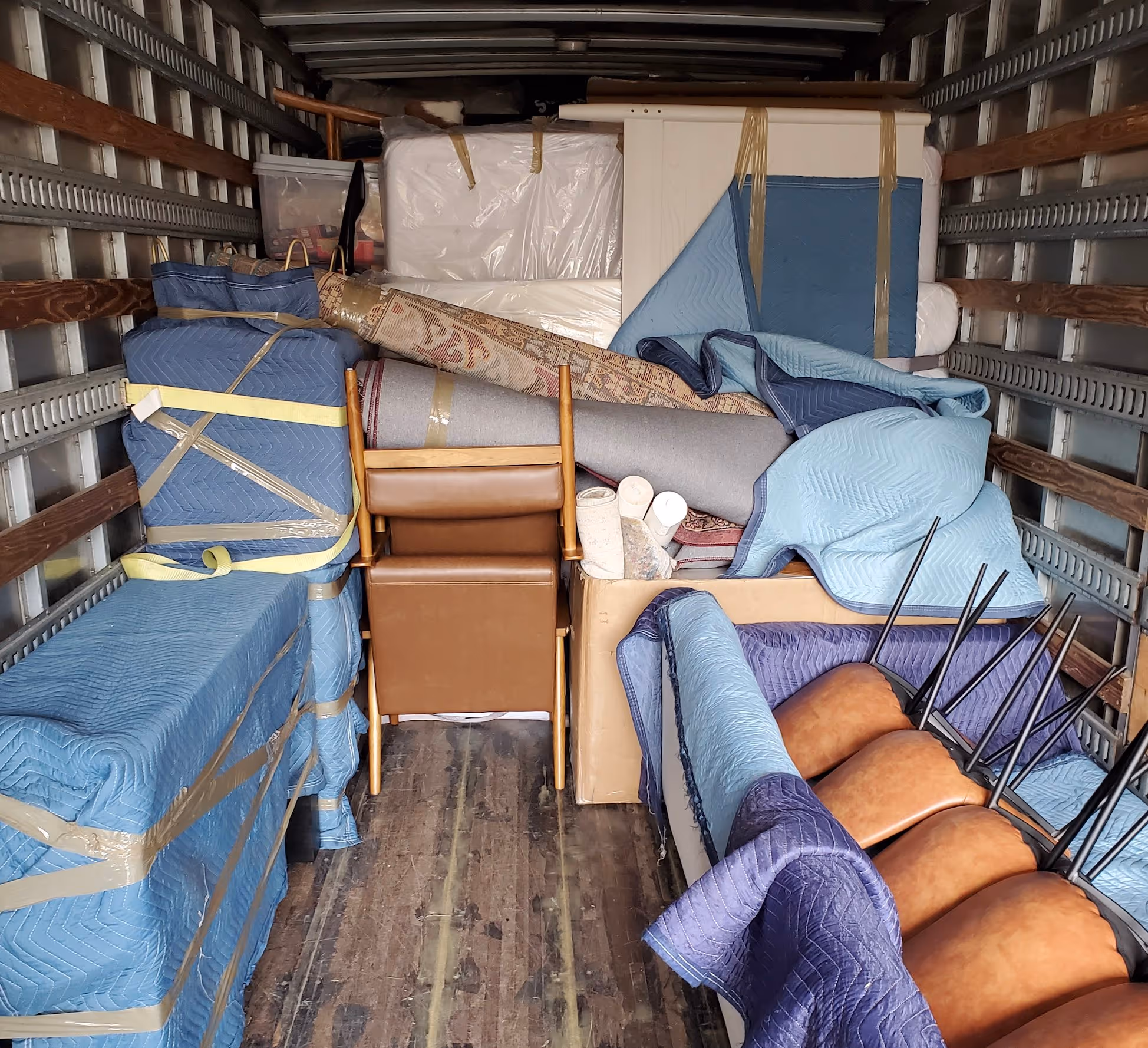Moving truck interior with furniture (chairs, bed frame, rolled rugs) secured and protected with blue and lighter blue moving blankets, highlighting professional furniture packing and moving services.