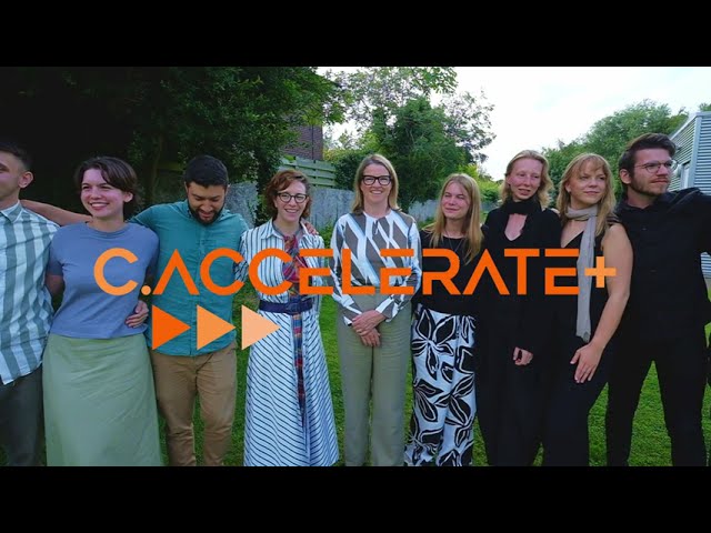 Group of eight diverse people standing closely together outdoors on grass, smiling with trees and a building in the background, overlaid with orange text reading 'C-ACCELERATE+'.