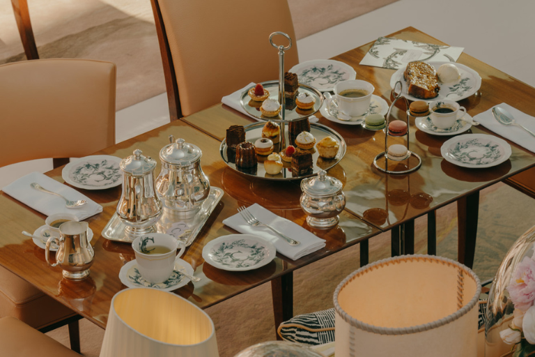 Elegant wooden tea table with cups, teapot, two-tier pastry tray and macaroon stand surrounded by beige chairs.