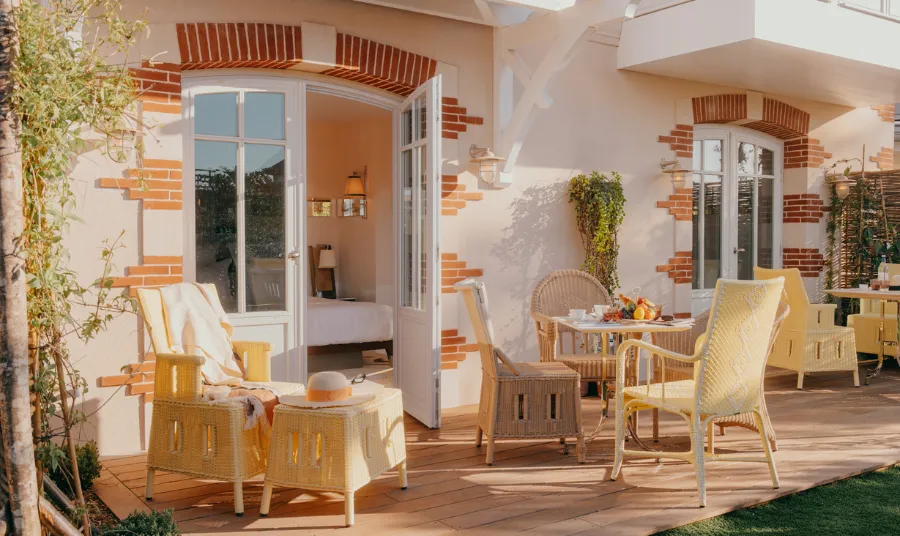 Sunny terrace with light yellow rattan chairs, table with fruits and drinks, open glass doors leading to a bedroom.