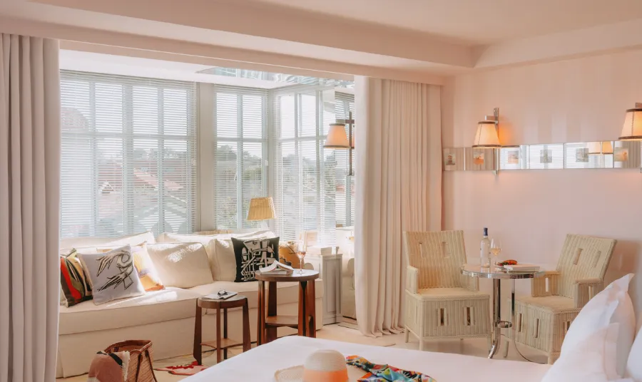 Bright living room with white sofa decorated with colorful cushions, two wicker armchairs around a round table with a bottle of wine.