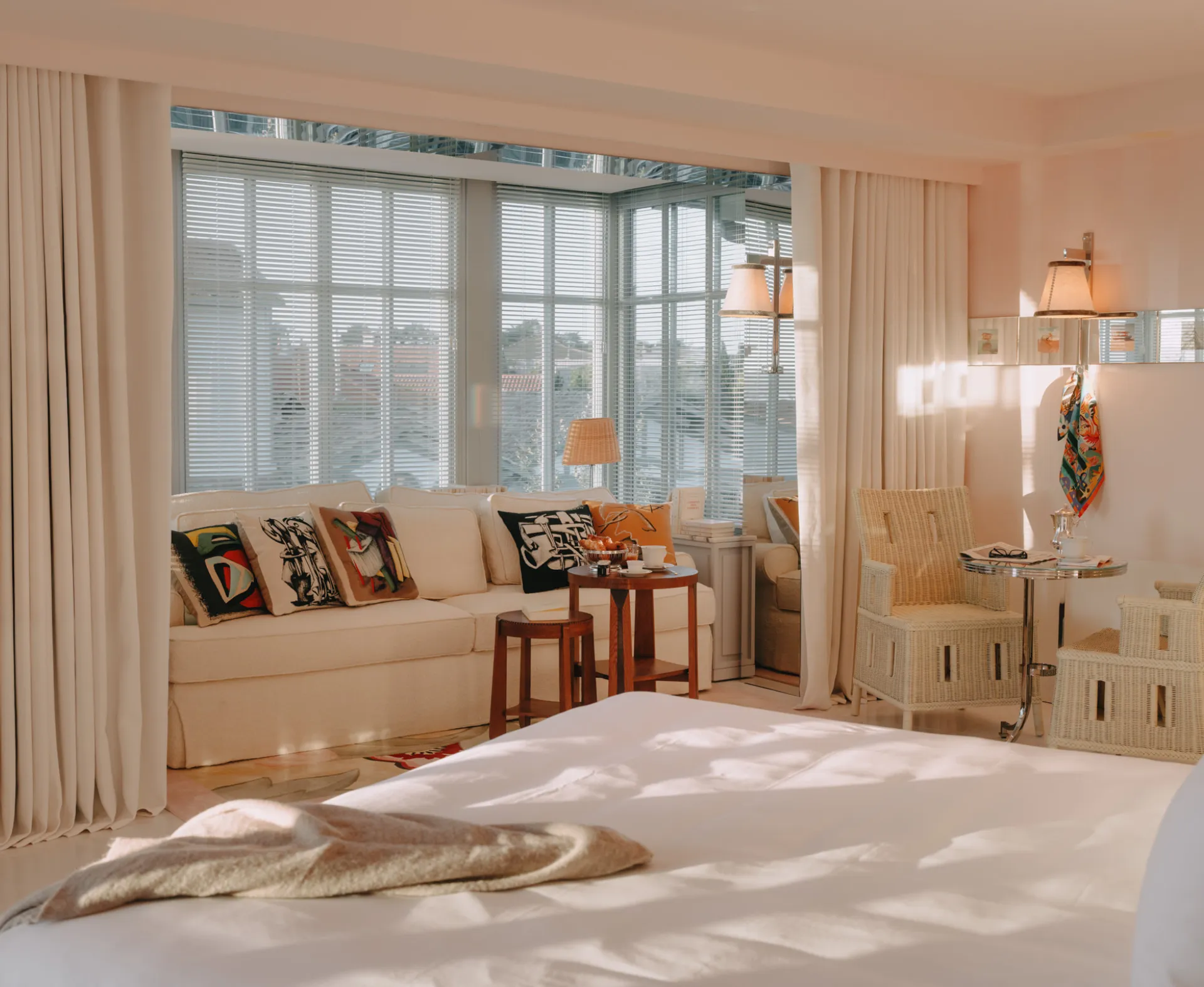 Bright room with a large white bed, a beige sofa with colorful cushions, a wooden table with lamps, and wicker armchairs near a window with blinds.