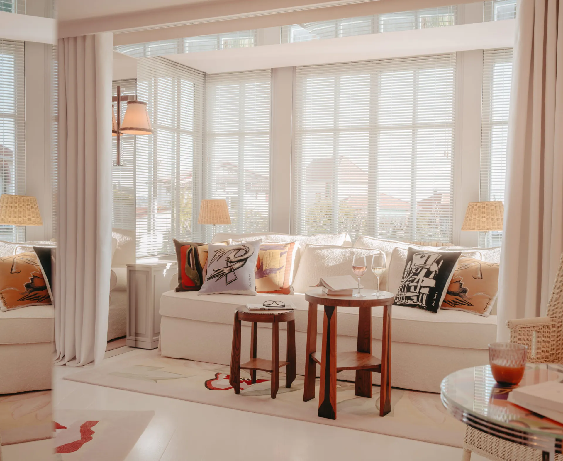 Bright living room with white sofa, decorative cushions, wooden tables with two glasses of wine, and large windows with Venetian blinds.