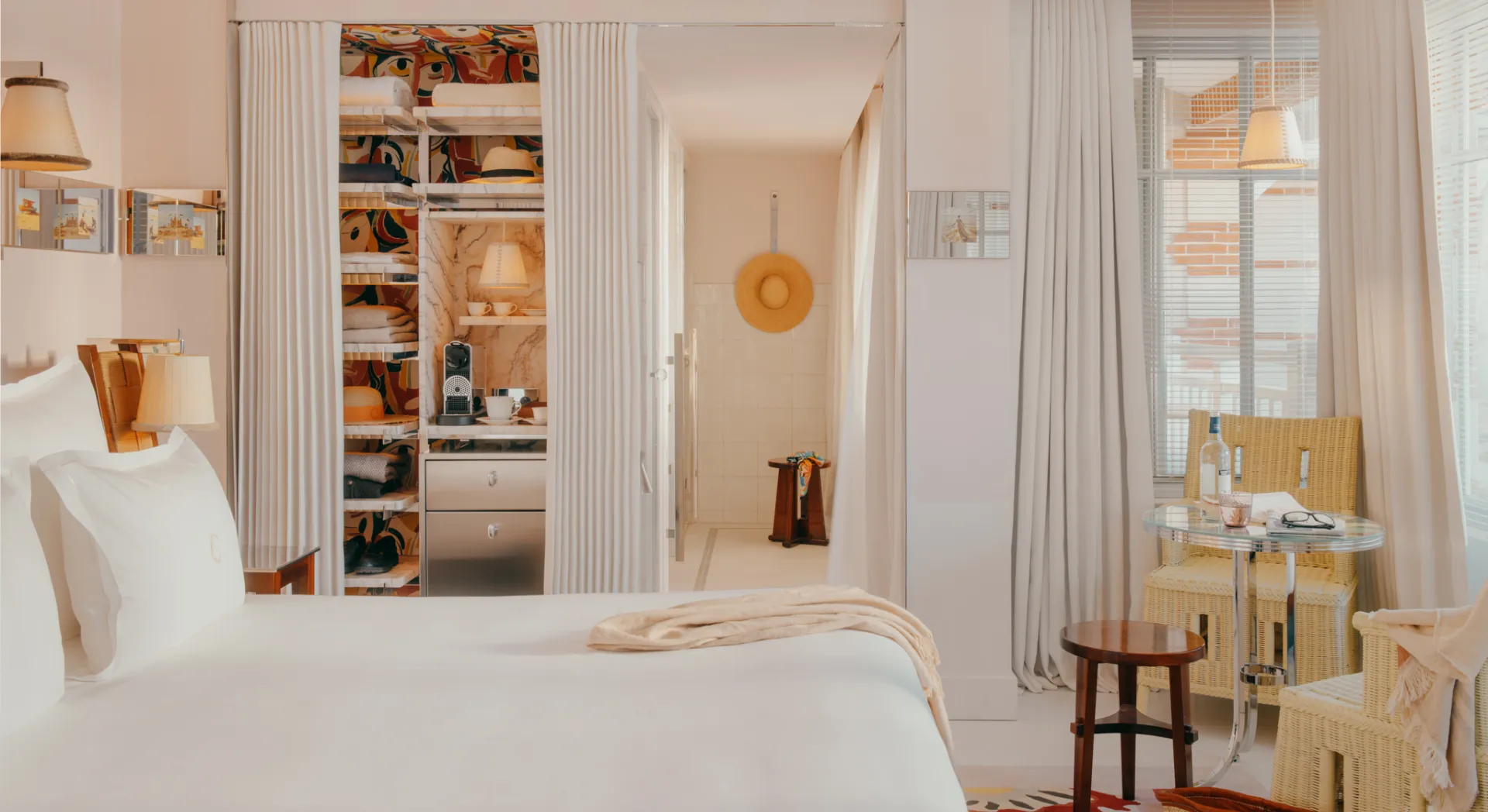 Bright hotel room with large white bed, wicker armchairs, glass table, and dressing area with coffee machine.