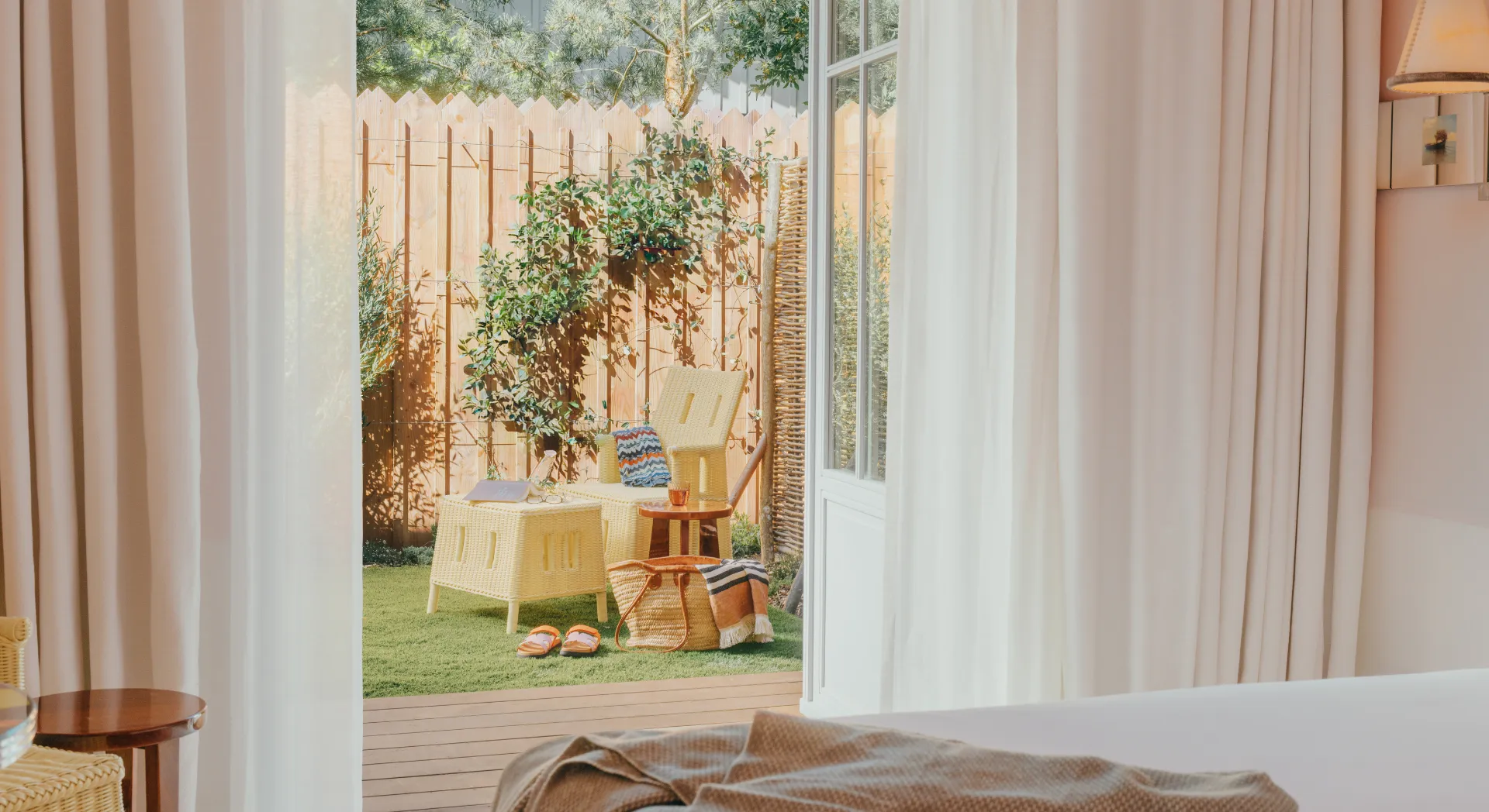 View from a bedroom to a terrace with wicker armchair, small table, basket and green lawn surrounded by a wooden fence.