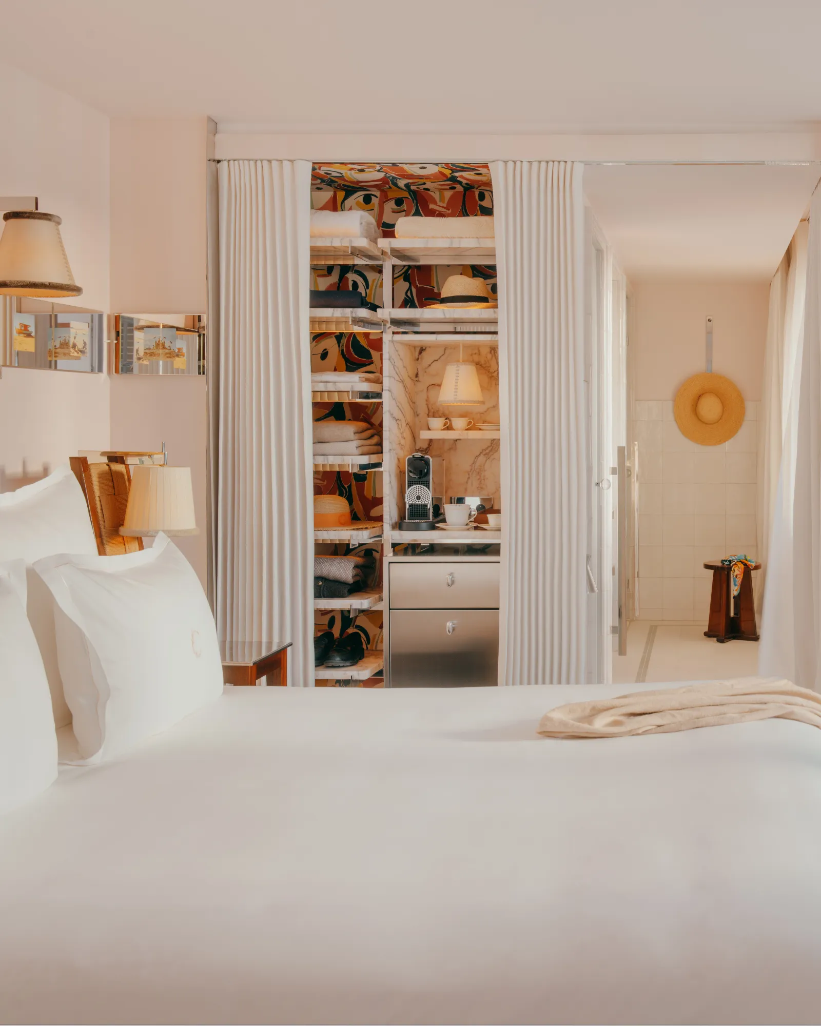 Stylish hotel room with large white bed, open shelves with clothes and accessories, coffee machine, and bright bathroom with a hanging hat.