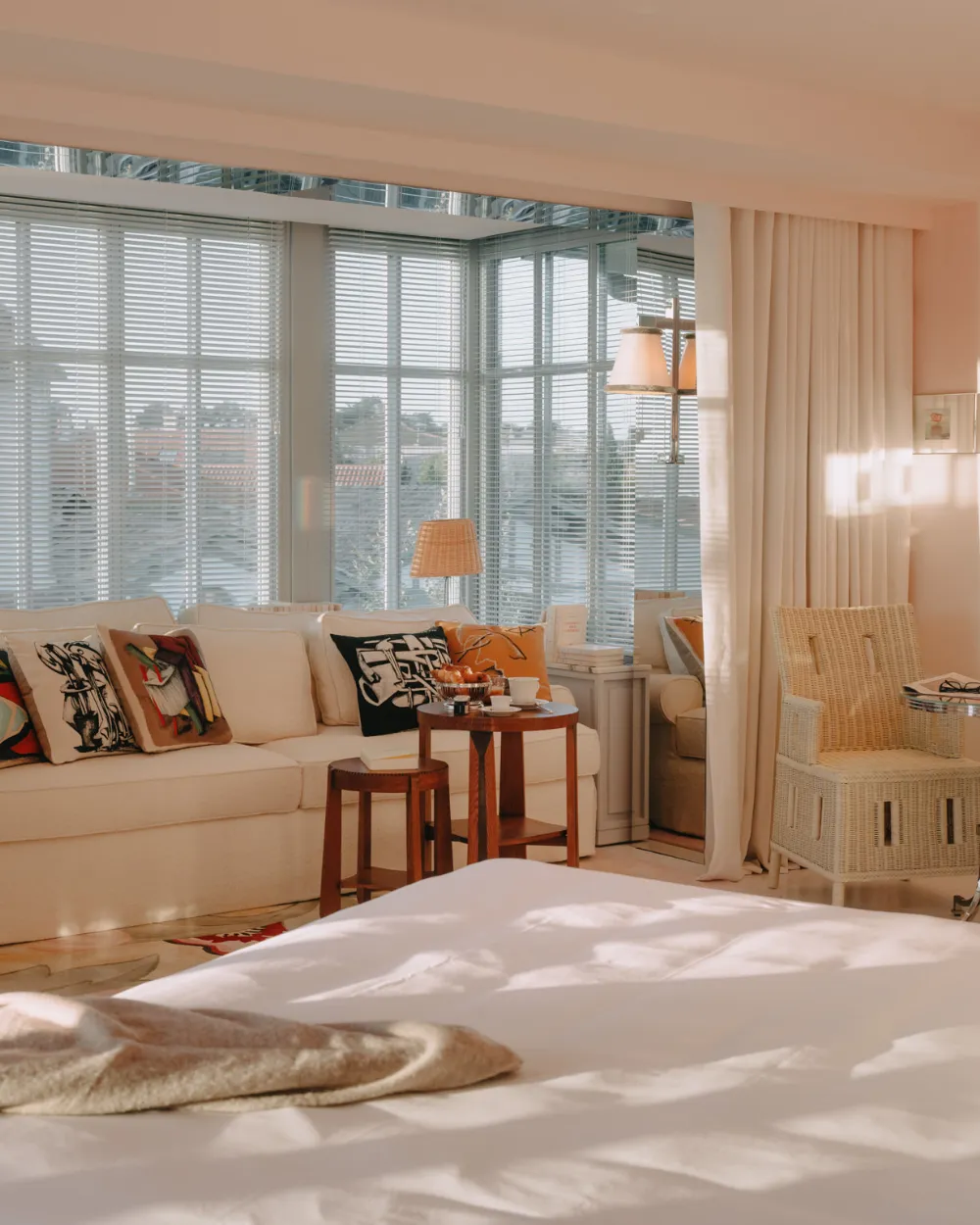 Bright bedroom with large white bed, beige sofa decorated with artistic cushions, small wooden table with cups, wicker lamp, and rattan chair near shuttered windows.