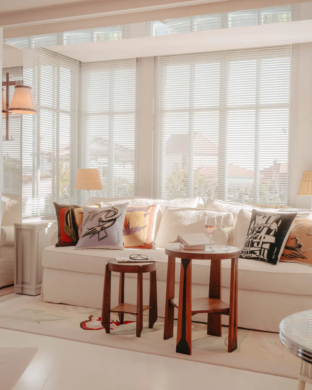 Bright living area with white sofa, decorative cushions, two wooden tables with wine glasses, lamps and large windows with white blinds.